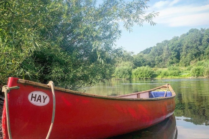 Red canoe labeled 'HAY' on a calm riverbank with trees and a clear blue sky.