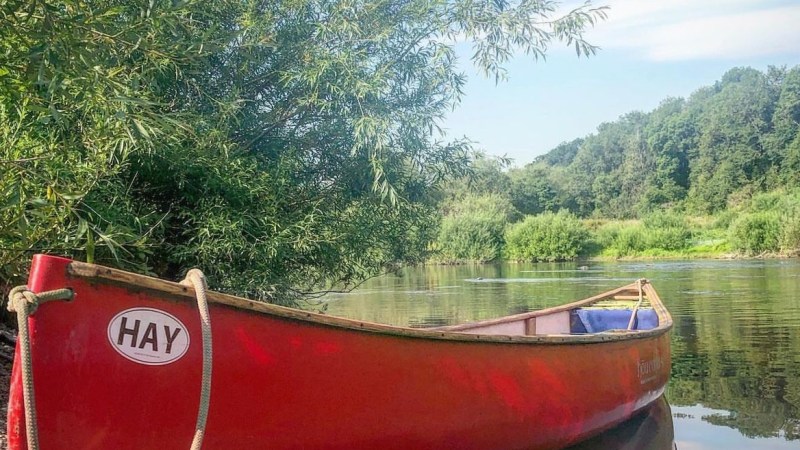 Red canoe labeled 'HAY' on a calm riverbank with trees and a clear blue sky.