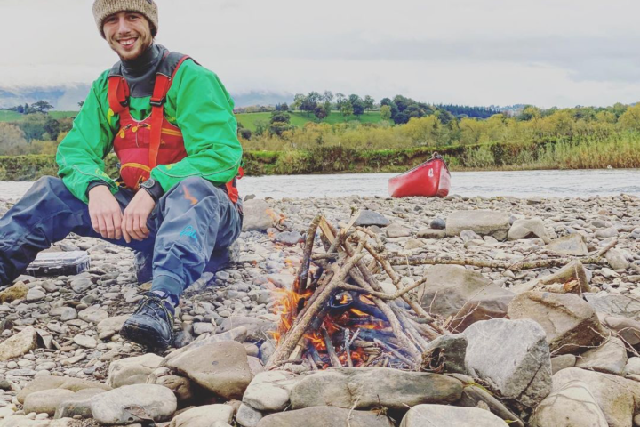 Man in green jacket sitting by a fire on a rocky shore; red canoe in the background.