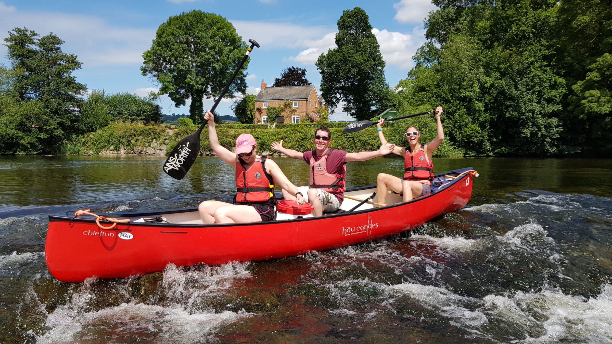 Three people in a red canoe paddling on a river, surrounded by trees and a house in the background.