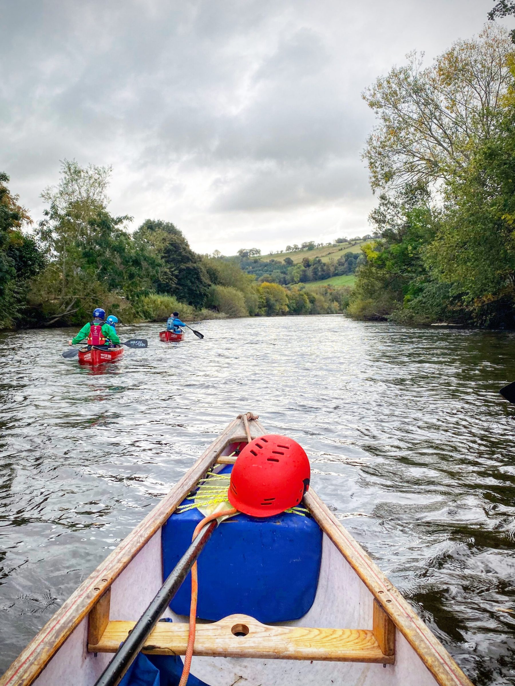 Canoes on a river with people paddling, surrounded by trees and cloudy sky.