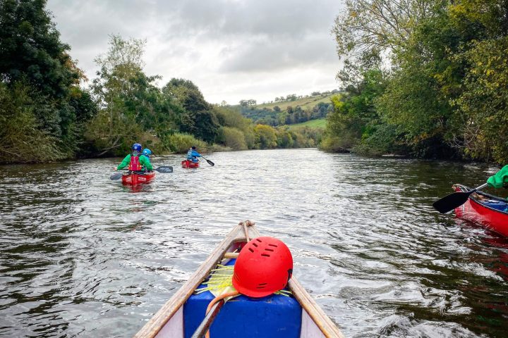 Canoes on a river with people paddling, surrounded by trees and cloudy sky.