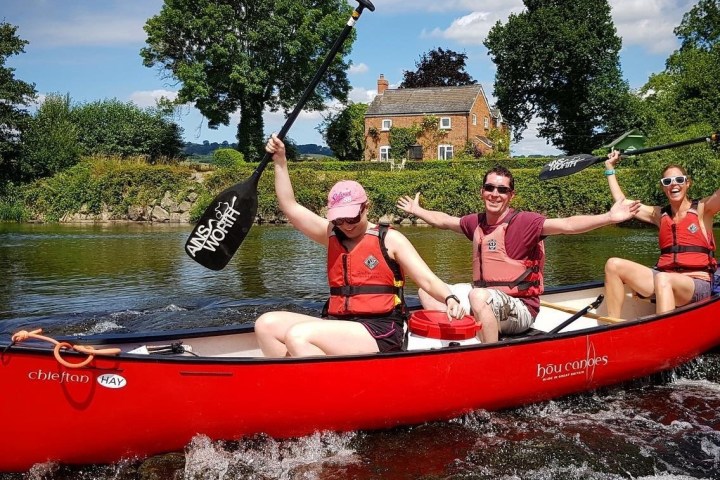 Three people in a red canoe on a river, holding paddles joyfully with a house in the background.