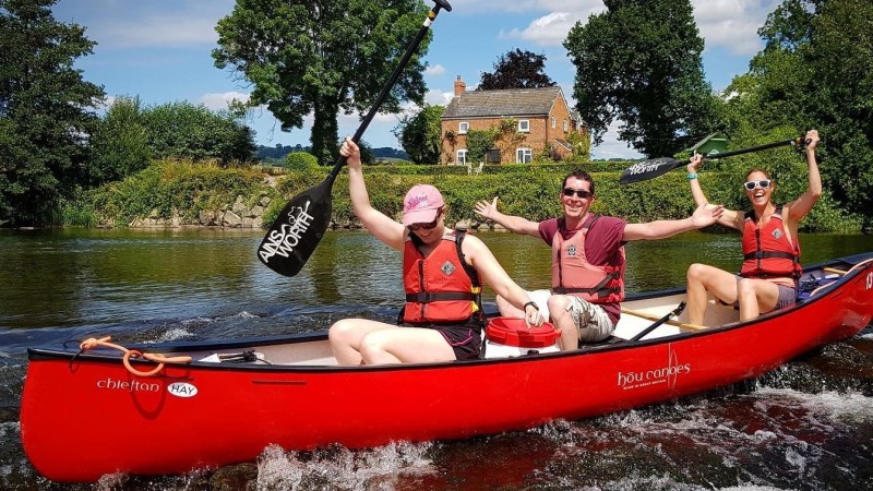 Three people in a red canoe on a river, holding paddles joyfully with a house in the background.