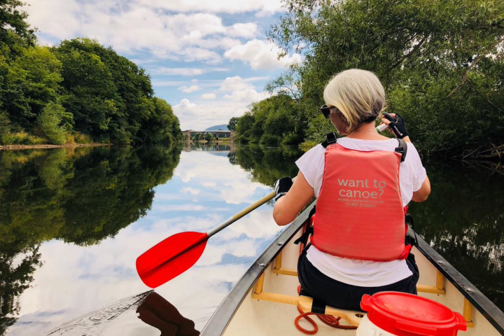 Person in red vest paddling a canoe on a calm river surrounded by trees under a partly cloudy sky.