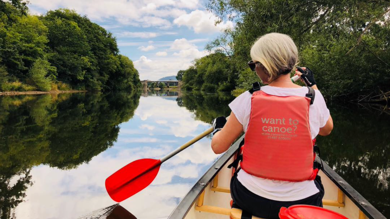 Person in red vest paddling a canoe on a calm river surrounded by trees under a partly cloudy sky.