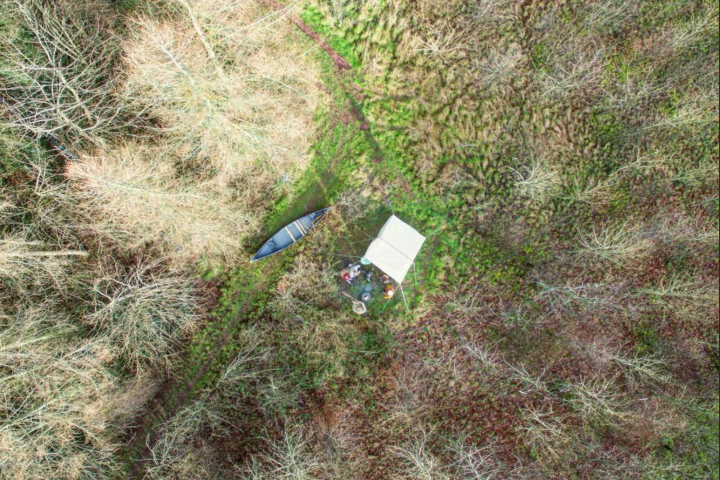 Aerial view of a tent and canoe in a forest clearing with bare trees.
