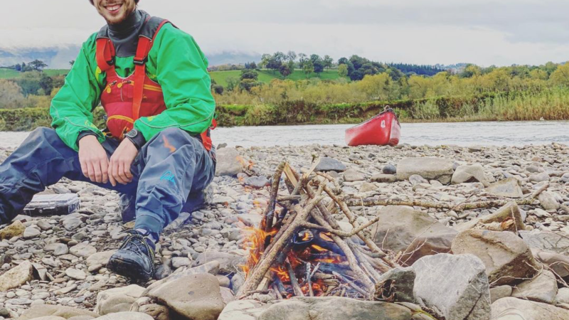 Person in outdoor gear sits by campfire on rocky shore with red canoe and river in background.