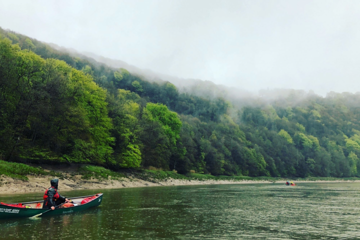 Person in a canoe on a misty river, with a forested hillside in the background.
