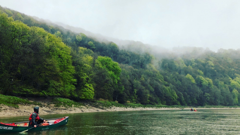 Person in a canoe on a misty river, with a forested hillside in the background.