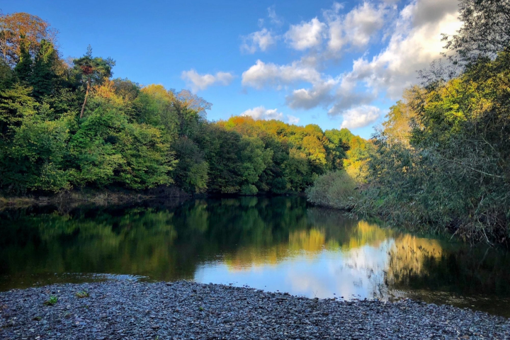 A calm river with tree reflections under a blue sky with clouds.