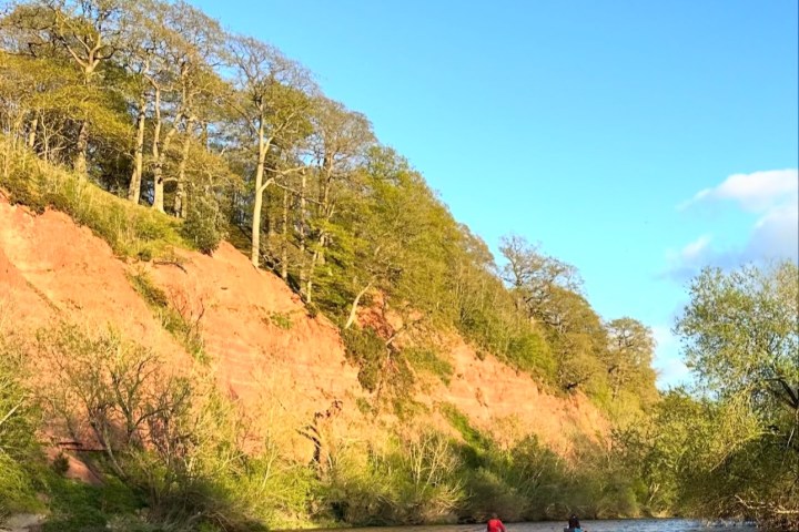 Two people kayaking on a river beside a rocky cliff with trees under a blue sky and moon.