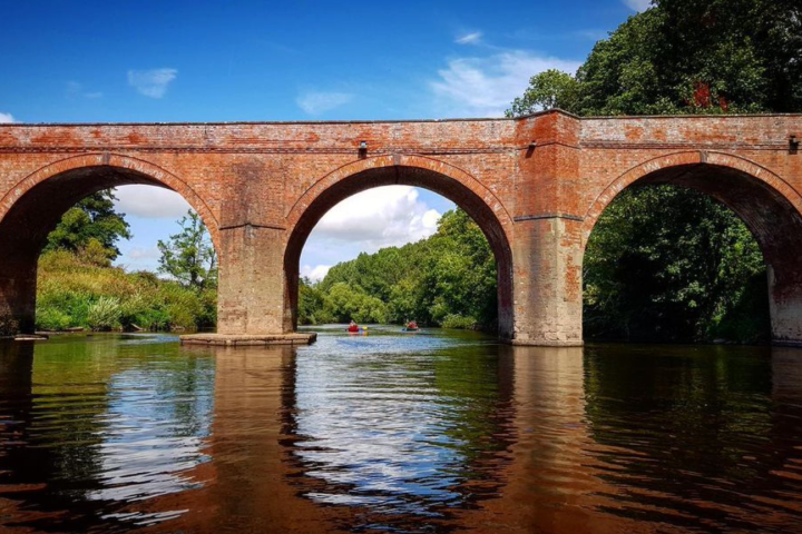 Three-arch red brick bridge over a calm river with lush green trees and a blue sky.