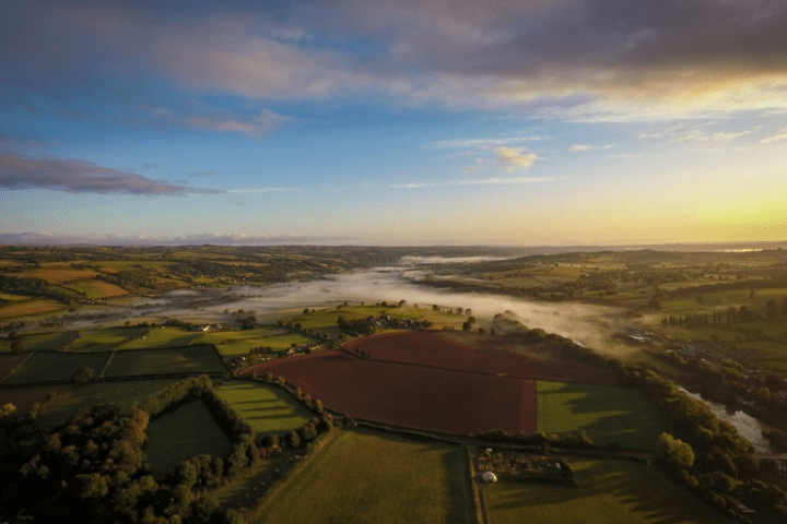 Aerial view of a misty rural landscape at sunrise with fields and trees.