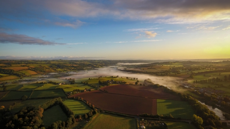 Aerial view of a misty rural landscape at sunrise with fields and trees.