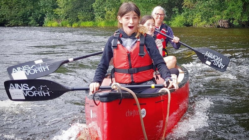 Three people in red life jackets paddling a red canoe on a river with forested banks.