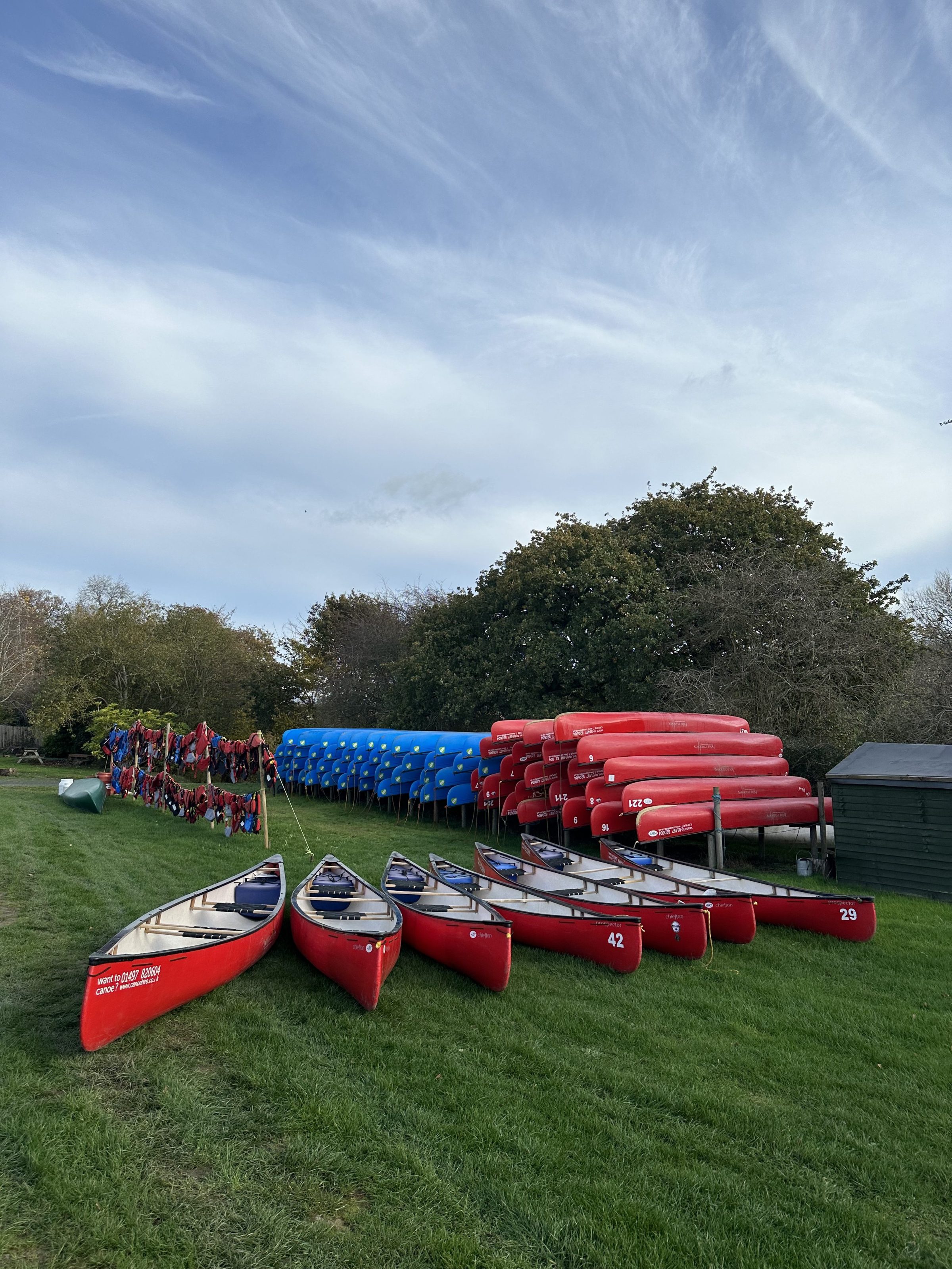 Red and blue canoes stacked on grass with trees and sky in background.