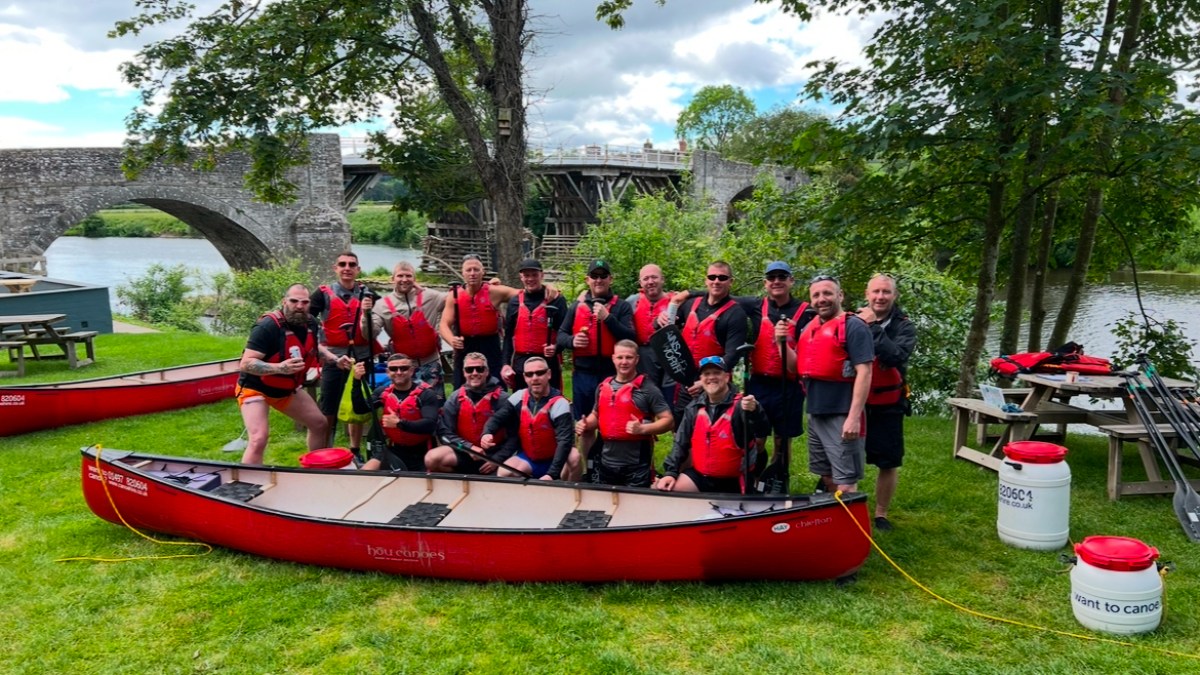 Group of people in red life vests pose with canoes near a river and bridge.
