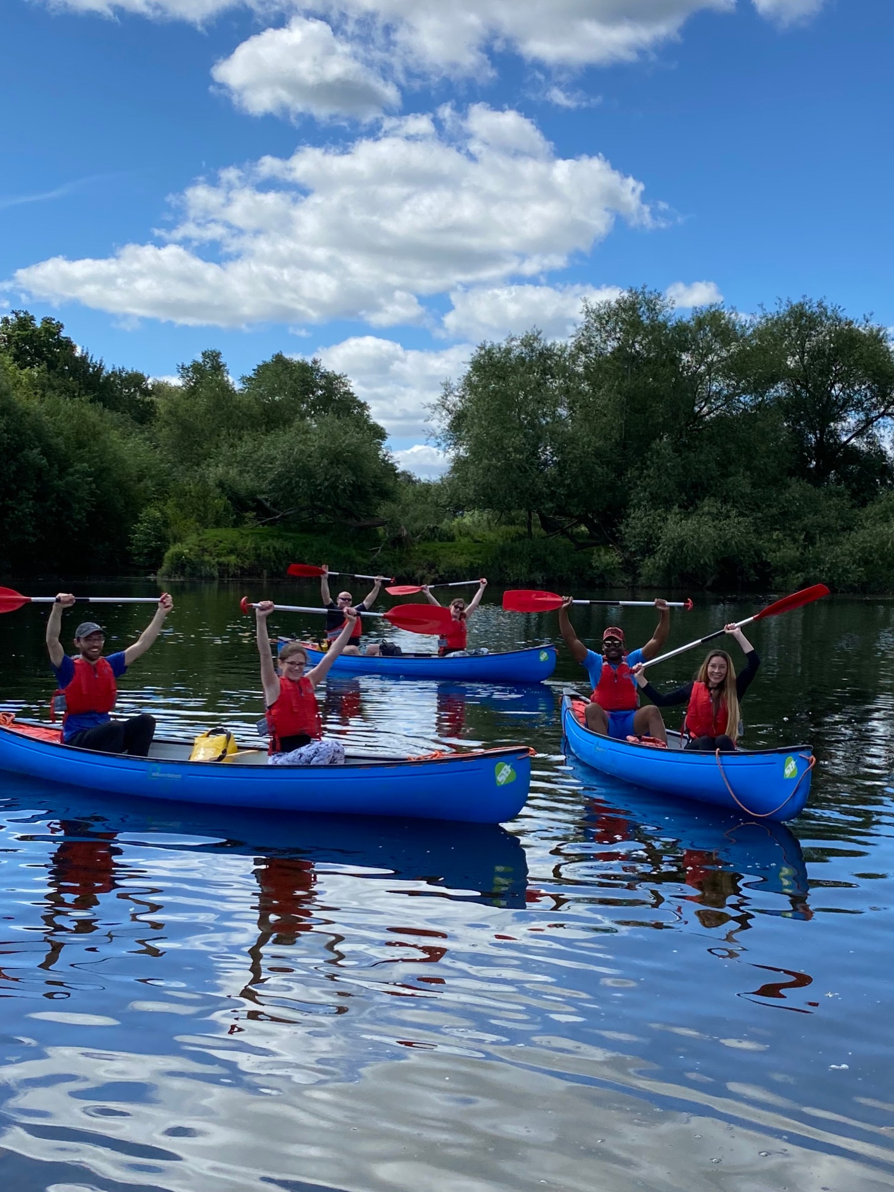 Five people in blue canoes with paddles on a lake, surrounded by trees under a blue sky with clouds.