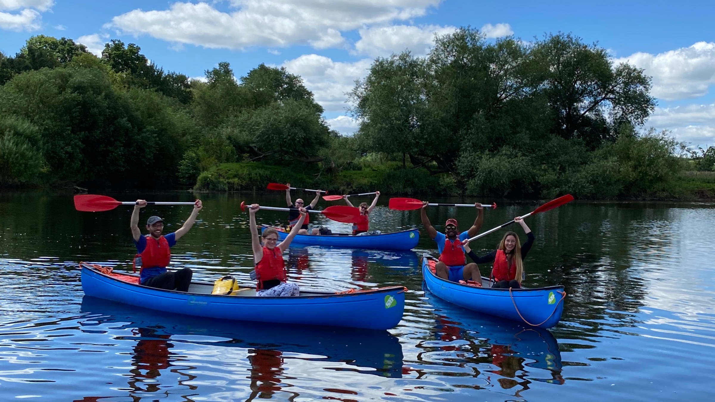 Five people in blue canoes with paddles on a lake, surrounded by trees under a blue sky with clouds.