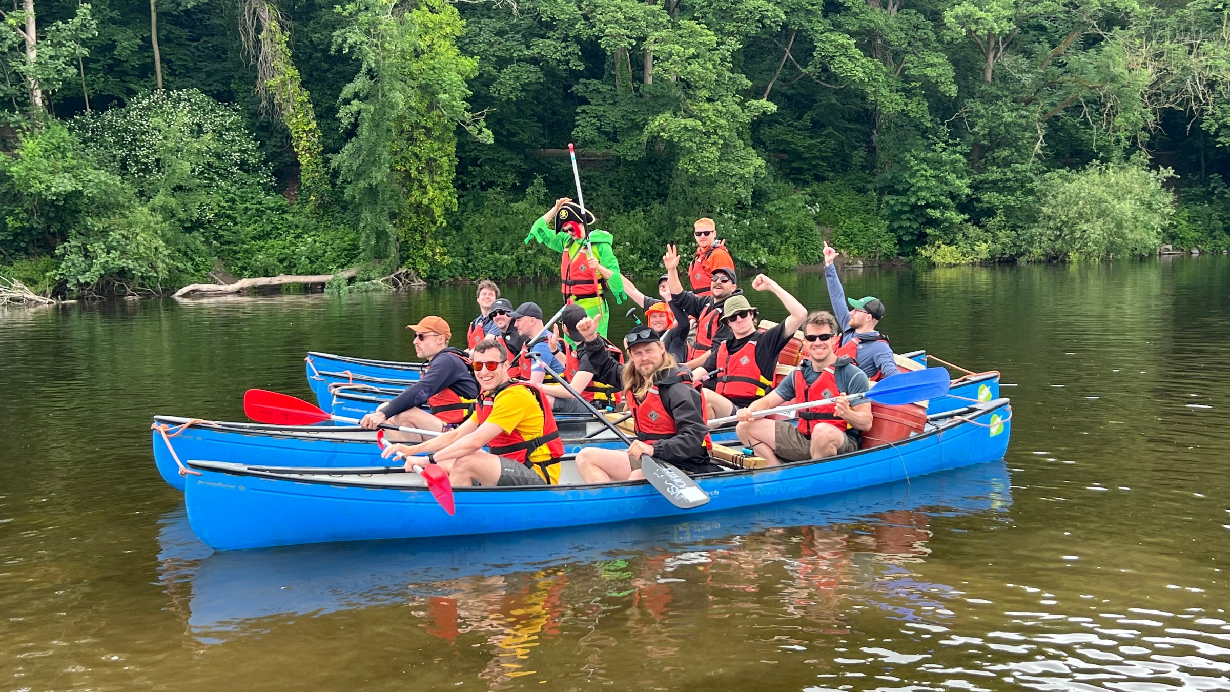 Group in blue canoes on a river, wearing life vests, surrounded by green forest.