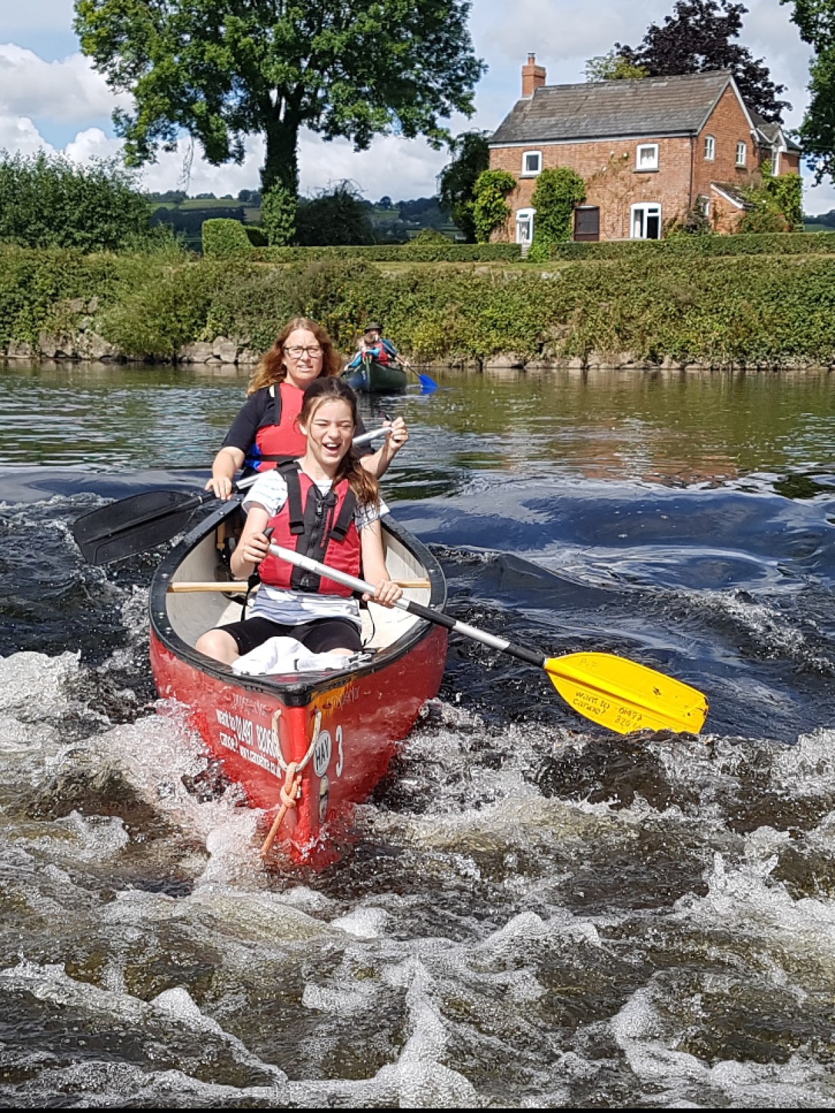 Two people canoeing in a river with a house and trees in the background.