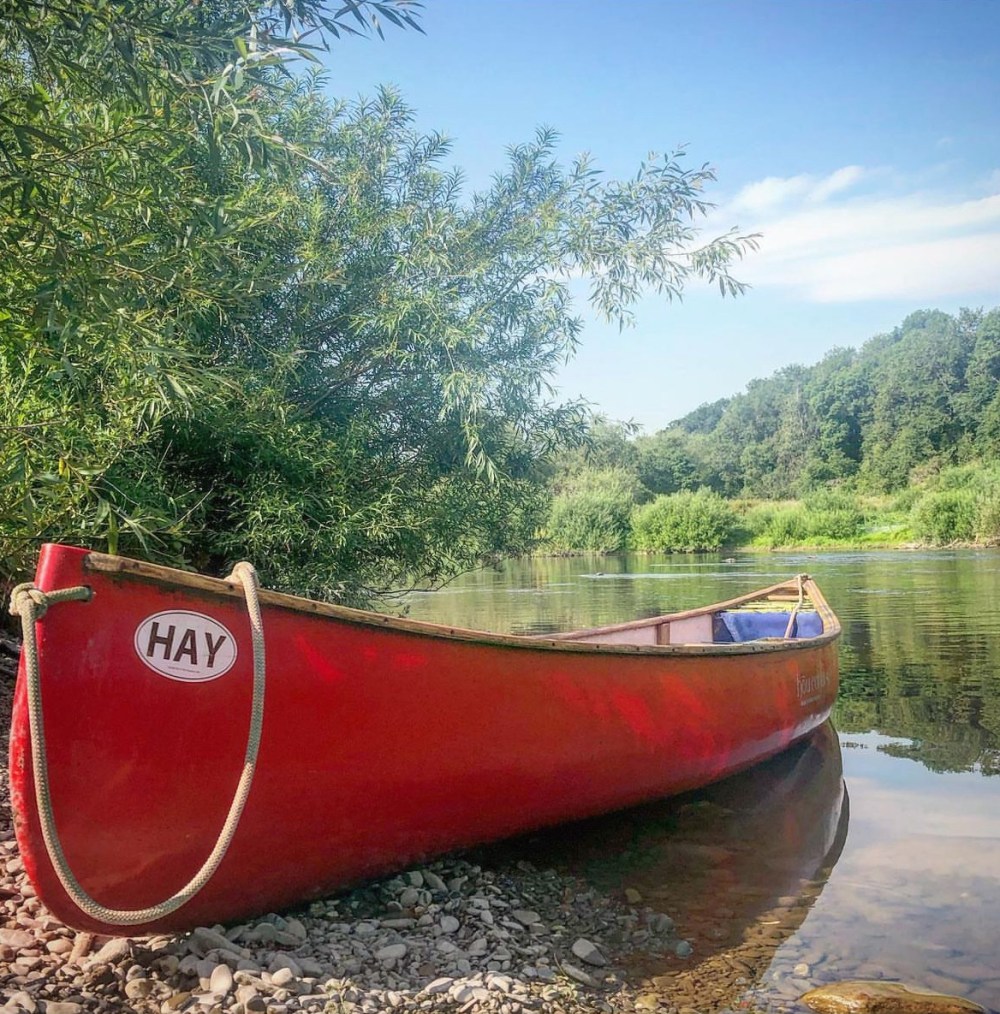 Red canoe on a rocky riverbank with trees and calm water under a clear blue sky.