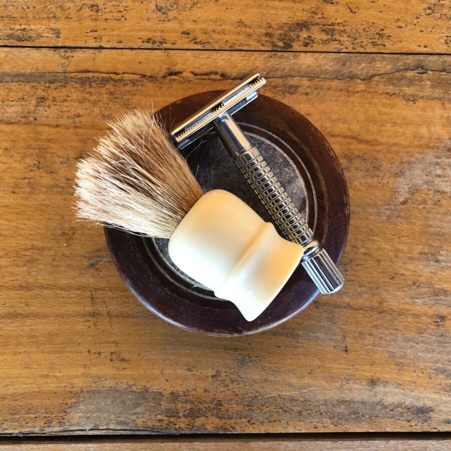 Shaving brush and razor on a wooden bowl on a rustic table.