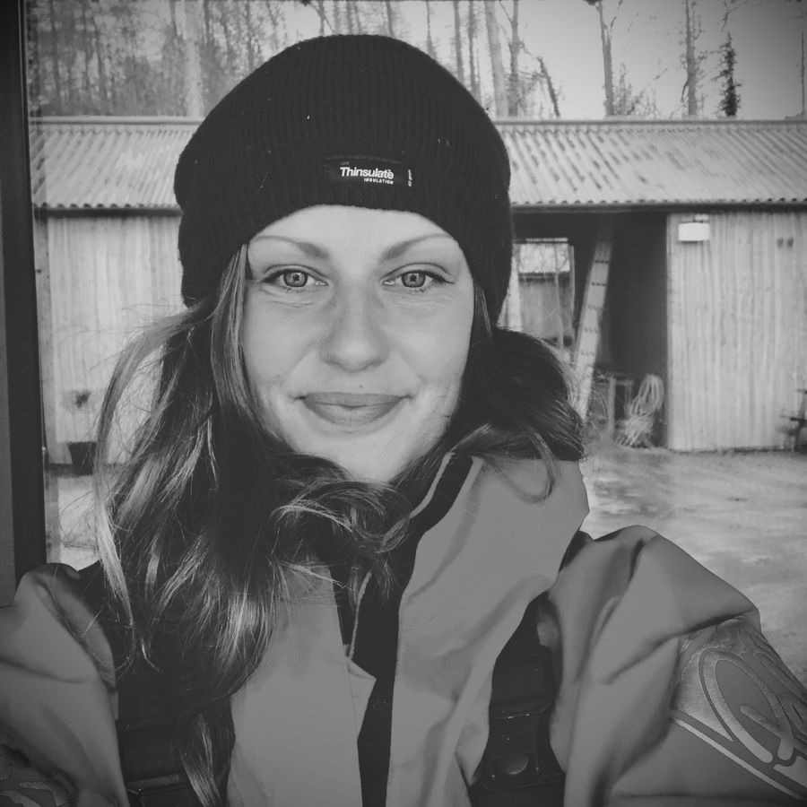 Person in black beanie and jacket smiles outdoors with wooden building background.