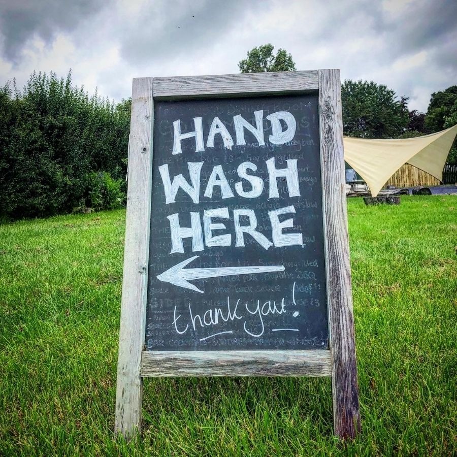 Chalkboard sign on grass reads: 'HAND WASH HERE' with an arrow pointing left.