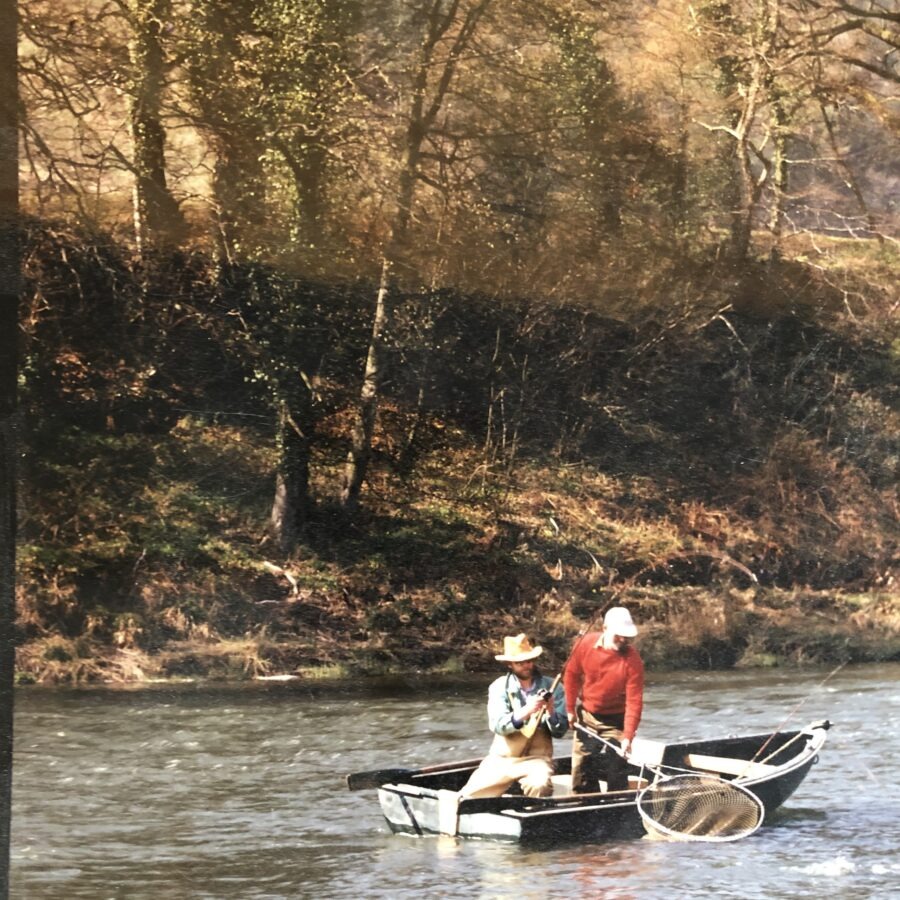 Two people fishing on a river in a small boat near a wooded riverbank.