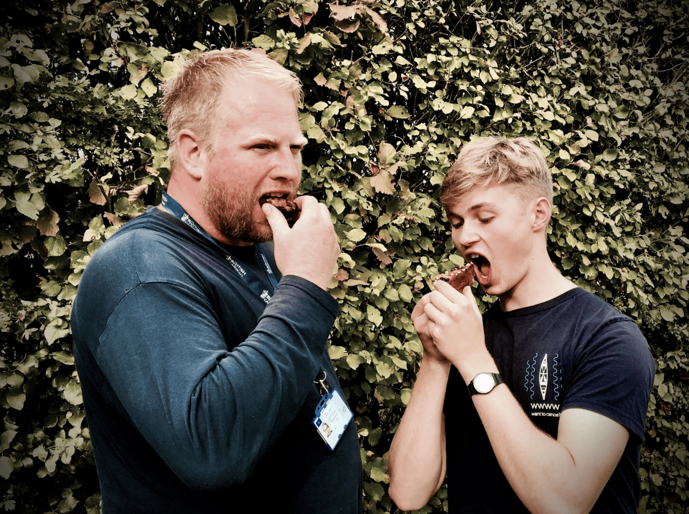 Two people eating chocolate brownies outdoors against a leafy background.