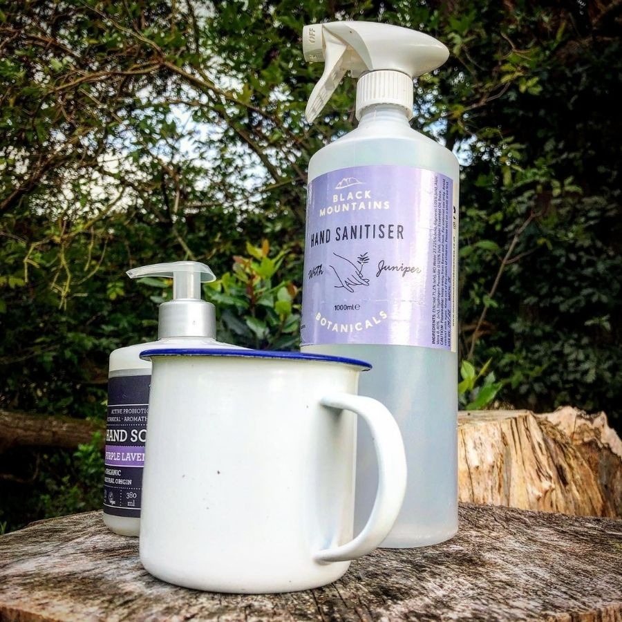Hand sanitiser, soap, and mug on tree stump, with greenery in background.