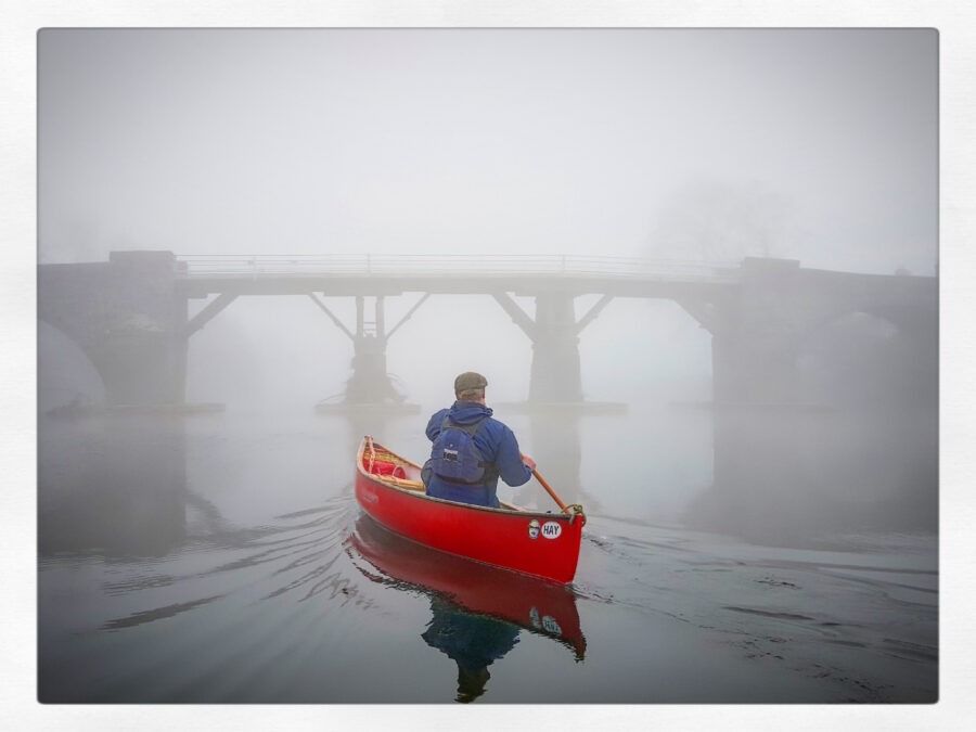 Person paddling a red canoe towards a foggy bridge over calm water.