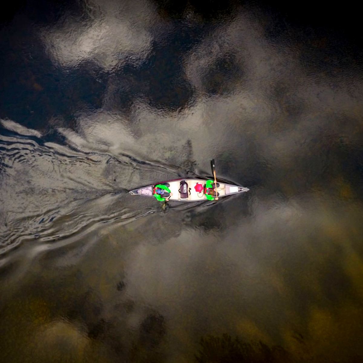 Aerial view of people in a canoe paddling on a reflective, cloudy water surface.