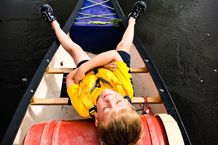 Child in yellow shirt relaxing in a canoe on water with eyes closed.