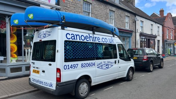 White van with blue canoes on top, parked on a street with colorful bunting overhead.